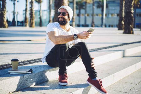 Foto de Chico hipster alegre en traje de moda sonriendo mientras descansa en el entorno urbano en la ciudad utilizando el dispositivo de teléfono celular en el ocio, feliz blogger masculino de Oriente Medio en gafas de sol disfrutando de un estilo de vida de viaje - Imagen libre de derechos