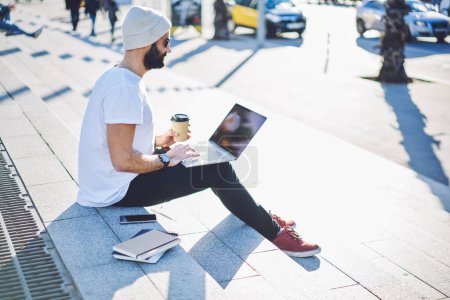 Nahöstlicher männlicher digitaler Nomade mit Coffee to go, der Projektinformationen überprüft, während er Software im städtischen Umfeld entwickelt, kompetenter Hipster, der Webpublikationen liest, während er soziale Medien vernetzt