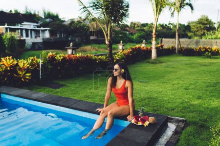 Téléchargez les photos : Belle jeune femme en maillot de bain rouge assise sur le bord de la piscine et obtenir les pieds mouillés tout en ayant des vacances à la station - en image libre de droit