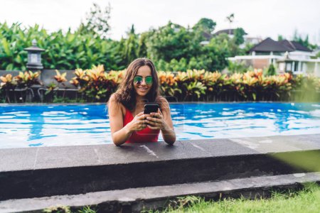 Foto de Joven morena alegre en traje de baño y gafas de sol navegando por el teléfono inteligente y sonriendo mientras pasa tiempo en la piscina en un día soleado en un resort tropical - Imagen libre de derechos