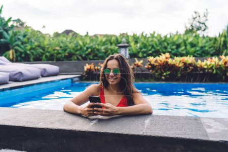 Téléchargez les photos : Femme gaie en maillots de bain et lunettes de soleil navigation smartphone tout en se reposant et se penchant sur le bord de la piscine dans une journée d'été ensoleillée - en image libre de droit