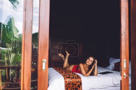 Téléchargez les photos : Délicieuse voyageuse portant un maillot de bain couché sur un lit confortable dans une chambre d'hôtel moderne et profitant de vacances d'été tout en regardant la caméra - en image libre de droit