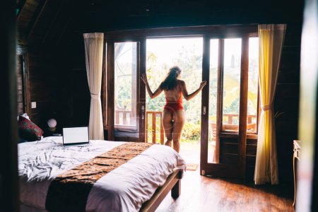 Foto de Vista posterior de la hembra delgada en traje de cuerpo rojo admirando la naturaleza y respirando aire fresco mientras está de pie en la puerta del dormitorio al amanecer - Imagen libre de derechos