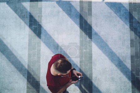 Téléchargez les photos : D'en haut de l'homme athlétique de culture en t-shirt rouge naviguant smartphone sur la chaussée pendant la pause et écouter de la musique à travers des écouteurs - en image libre de droit