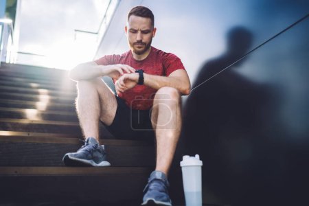 Téléchargez les photos : Angle bas de l'homme réfléchi en baskets regardant smartwatch après avoir fait de l'exercice de jogging sur la rue dans la journée ensoleillée d'été - en image libre de droit