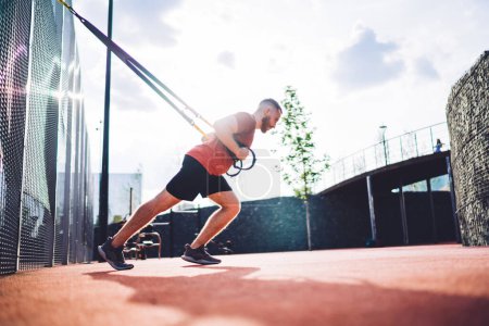 Téléchargez les photos : Vue latérale d'un sportif musclé faisant de l'exercice sur les sangles TRX et regardant ailleurs pendant l'entraînement le matin sur un terrain de sport - en image libre de droit