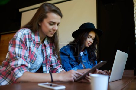 Téléchargez les photos : Deux étudiantes en tenue décontractée et chapeau assis à table avec ordinateur portable tout en travaillant ensemble dans l'auditorium universitaire - en image libre de droit