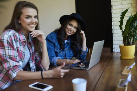 Téléchargez les photos : Jeunes femmes gaies en vêtements décontractés et chapeau assis à table avec ordinateur portable et téléphone portable tout en travaillant ensemble dans un café moderne - en image libre de droit