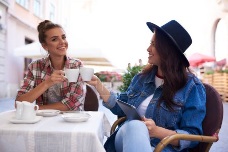 Téléchargez les photos : Femme positive avec tablette assise à table dans un café extérieur et boire du café avec un ami assis sur une chaise et souriant largement - en image libre de droit