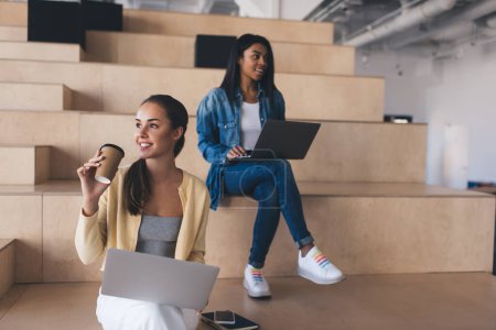 Photo for Multiracial businesswomen using laptops while working. Concept of modern successful women. Young european and black girls on wooden benches in coworking office space. Caucasian lady with coffee - Royalty Free Image