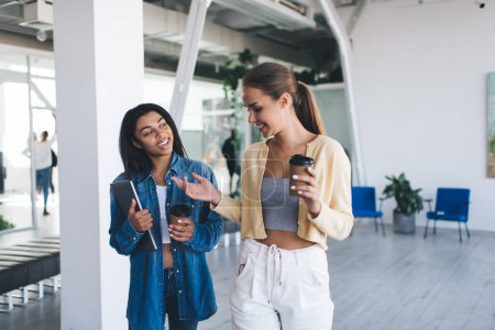 Photo for Multiracial businesswomen going and talking in open space office. Concept of modern successful women. Idea of teamwork. Young smiling european and black girls with digital devices and coffee - Royalty Free Image