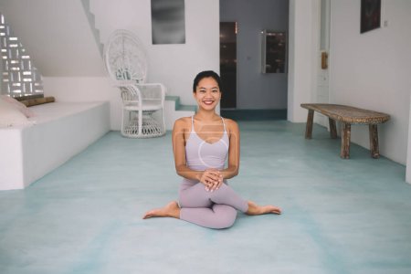 Asian woman practicing yoga in Lotus pose at spacious home. Concept of harmony and mental health. Young smiling beautiful girl wearing sportswear and barefoot sitting on floor and looking at camera