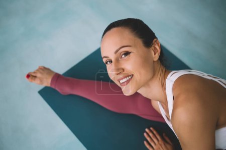 Partial image of caucasian girl practicing yoga in Seated wide angle straddle pose at home. Concept of healthy lifestyle. Young smiling woman wear sportswear sit on fitness mat and look at camera