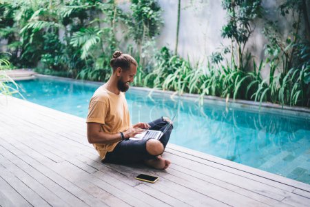 Sonriente freelancer masculino navegando contenido de video mientras hace networking en redes sociales usando tecnología netbook en la terraza de la piscina, chico hipster feliz blogueando en línea a través de un portátil disfrutando del estilo de vida nómada digital