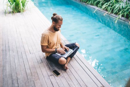 Millennial männlich mit Laptop-Computer arbeitet an Online-Projekt Browsing-Websites in der Nähe des Pools in der Villa, Software-Programmierer kommuniziert in sozialen Netzwerken, während die Erstellung von Web-Design über Netbook-App