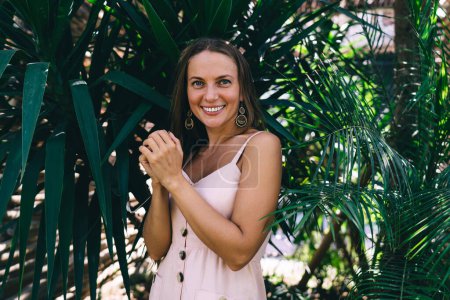 Half length portrait of cheerful female tourist dressed in casual wear smiling at camera during daytime leisure for exploring tropical environment with evergreen plants, happy woman on vacations