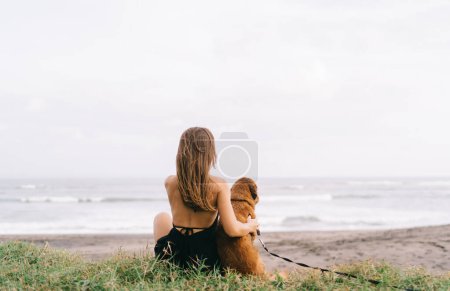 Rückansicht der kaukasischen Frau, die mit einem entzückenden Mischlingshund ihre Freizeit am Strand verbringt, eine unbeschwerte Frau, die sich an der Küste erholt, mit einem niedlichen Hundehündchen, das die Freundschaft im Freien genießt