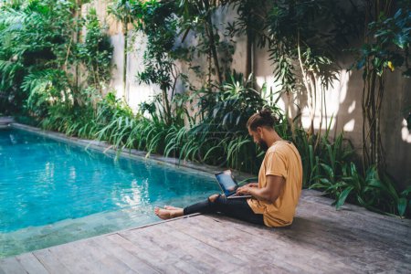 Skilled male copywriter keeping freelance digital nomad lifestyle - doing distance job at pool terrace of Bali, travel blogger creating content text using laptop for messaging in social media