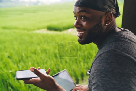 Fröhliche männliche Freiberufler 20er Jahre mit modernem Laptop und Smartphone-Gerät, die im Freien lächeln, fröhliche afroamerikanische Reiseblogger mit digitaler Mobiltechnologie, die sich über Reisfelder freuen