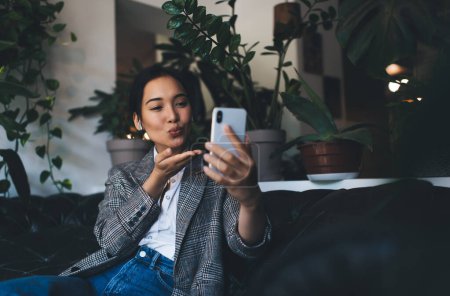 Téléchargez les photos : Blogueur web millénaire dans les écouteurs envoyer baiser d'air lors de la conférence vlog en ligne dans les réseaux sociaux, Utilisateur féminin asiatique dans les écouteurs se connectant à 4g sans fil pour faire des photos selfie - en image libre de droit