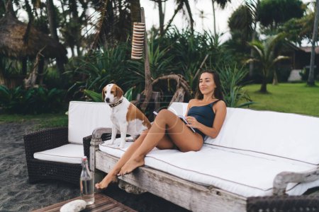 Caucaisan woman in swimwear resting in lounge zone with purebred dog in collar enjoying weekend leisure for reading interesting literature novel, young female with best seller book and fluffy friend