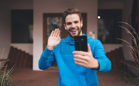 Téléchargez les photos : Millennial hipster guy dans les écouteurs profiter de l'appel vidéo pendant les loisirs en ville, gai blogueur masculin dans les écouteurs électroniques créer du contenu selfie en cliquant sur les photos sur la caméra web - en image libre de droit