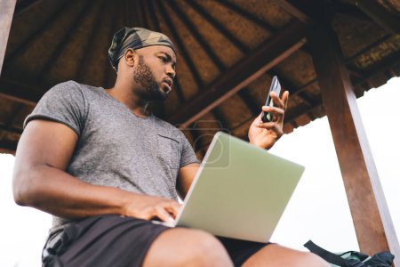 Afroamerikaner nutzt Mobiltelefon-Gerät zum Teilen von Mediendateien auf Laptop-Computer, der sich mit 4g Wireless und Bluetooth verbindet, Millennial-Grafikdesigner mit Netbook-Chat per Handy-App