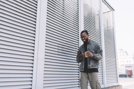 Millennial Black Tourist lächelnd in der Stadt mit Handy-Anwendung zur Ortung von GPS-Standort während der Reise zu Fuß, fröhliche Hipster Kerl in optischen Brillen genießen Online-Netzwerk und Chat