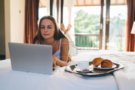 Millennial-Frau im seidenen Nachthemd mit Laptop-Anwendung zur Buchung von Hotelzimmer-Service im Online-Modus, kaukasischer digitaler Nomade im Chat in sozialen Netzwerken über Netbook-Technologie zu Hause