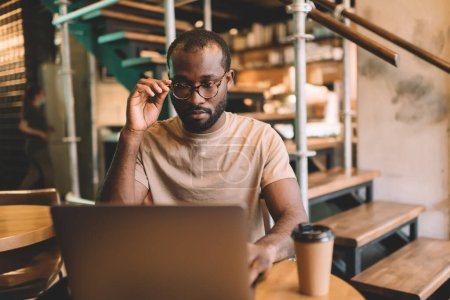 Qualifizierter Afroamerikaner in klassischer Brille surft Webinar-Video auf modernem Netbook-Gerät mit WIFI-Internetverbindung im Coworking Space, Millennial-Softwareentwickler macht Banking