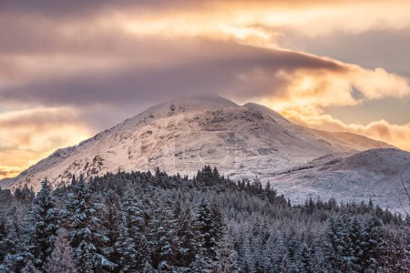 Der schneebedeckte Gipfel des Stob a 'Choire Mheadhoin über einem Tannenwald bei Sonnenuntergang in den schottischen Highlands, vom Laggan Dam aus gesehen