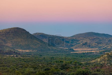 Ein rosafarbener Himmel leuchtet über Hügeln und üppiger Vegetation im Pilanesberg Nationalpark, der in einem uralten Vulkankrater in Südafrika liegt.