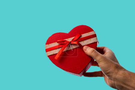 Téléchargez les photos : Man's hand giving a gift in a heart-shaped box with a bow, on a blue background. - en image libre de droit
