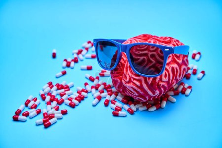 Red rubber brain with sunglasses on surrounded by white and red capsules on a blue surface. Concept of relaxation.