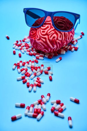 Cerebro rojo con gafas de sol y un montón de pastillas a su alrededor, encima de una superficie azul