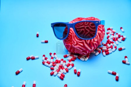 Cerebro rojo con gafas de sol y un montón de pastillas a su alrededor, encima de una superficie azul.