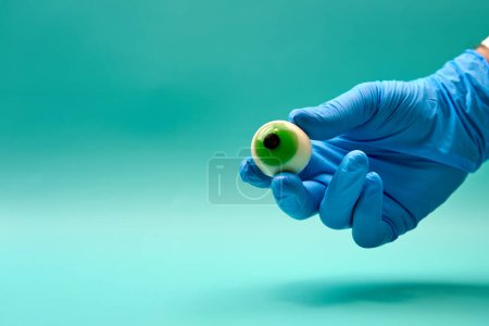 Foto de Mano de oftalmólogo irreconocible en guante de látex azul mostrando globo ocular artificial verde sobre fondo verde menta - Imagen libre de derechos