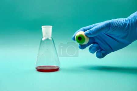 Nahaufnahme eines Titrationskolbens mit roter Flüssigkeit auf grüner Oberfläche und der Hand eines nicht erkennbaren Mediziners mit prothetischem Augapfel während eines Experiments im Labor