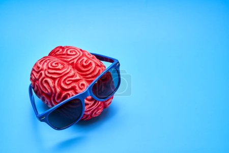 Rotes menschliches Gehirn aus Plastik mit blauer Sonnenbrille auf blauem Hintergrund. Trunkenheitskonzept.
