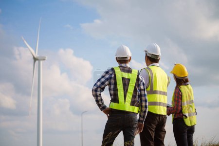 Back view of three engineers discussing and checking turbines on wind turbine farm. Renewable energy technology and sustainability.