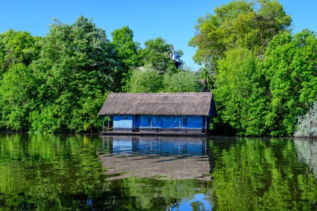 Téléchargez les photos : Paysage avec vieille maison bleue et beaucoup de grands arbres verts sur le rivage du lac Herastrau dans le parc King Michael I à Bucarest, Roumanie, dans une journée de printemps ensoleillée avec ciel bleu - en image libre de droit