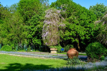 Landschaft mit grünen Bäumen, Blättern, Jahrgangsuhr und vielen kleinen Blumen an einem sonnigen Frühlingstag am Eingang zum Cismigiu-Garten (Gradina Cismigiu) in Bukarest, Rumänien
