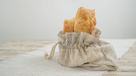 Artisanal ciabatta showcased in a sustainable linen bag, atop a wooden table adorned with a lovely tablecloth. Delicious Italian ciabatta displayed on a clean white backdrop.