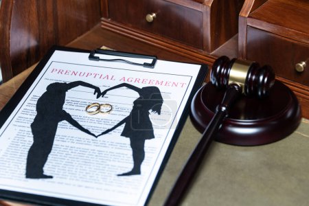 Photo for A clipboard holding a prenuptial agreement with a cut-out silhouette of a couple and wedding rings, next to a judges gavel - Royalty Free Image