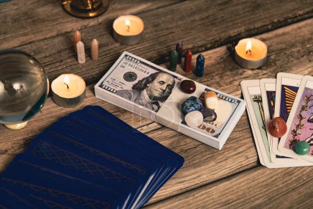 A tarot spread with The Fool card, hundred-dollar bills, and various crystals on a rustic wooden background with candles