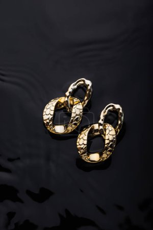 Téléchargez les photos : Boucles d'oreilles chaînes dorées sur fond d'eau noire. Bijoux concept de photographie - en image libre de droit