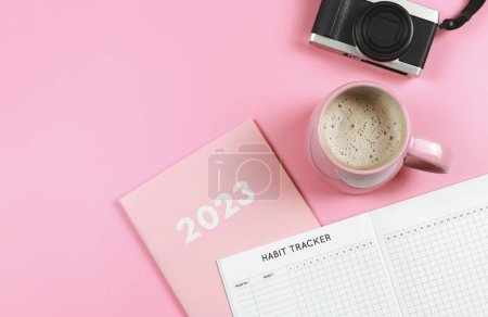 Top view or flat lay of habit tracker book on pink diary or planner 2023, pink cup of coffee and digital camera on pink background with copy space.
