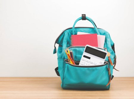 Foto de Vista frontal de la mochila verde con útiles escolares sobre mesa de madera y fondo blanco con espacio para copiar. - Imagen libre de derechos