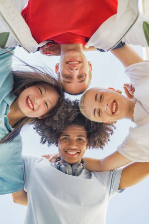 Vista de ángulo bajo de amigos jóvenes multiétnicos de moda abrazando, sonriendo y haciendo un círculo mirando a la cámara. Foto de alta calidad