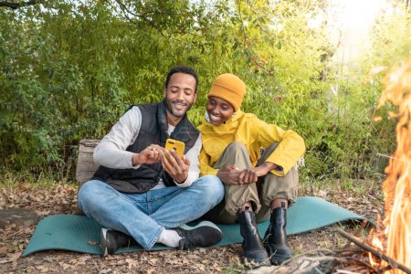 Téléchargez les photos : Jeune couple multiethnique assis autour du feu de camp regardant le téléphone mobile. Photo de haute qualité - en image libre de droit